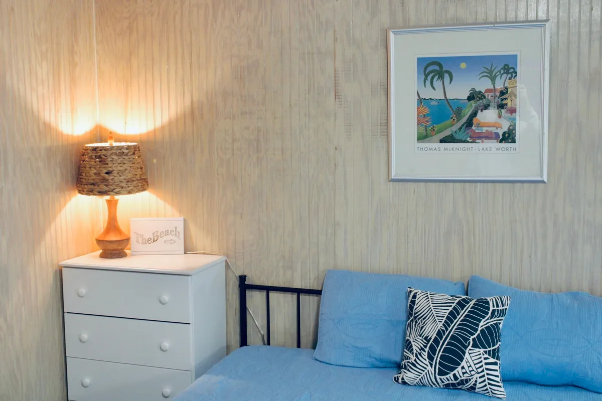 A cozy bed dressed in blue bedding sits next to a nightstand with a lamp, enhanced by colorful wall art and a decorative pillow. The space feels calm, welcoming, and ideal for unwinding after a day of coastal adventures.