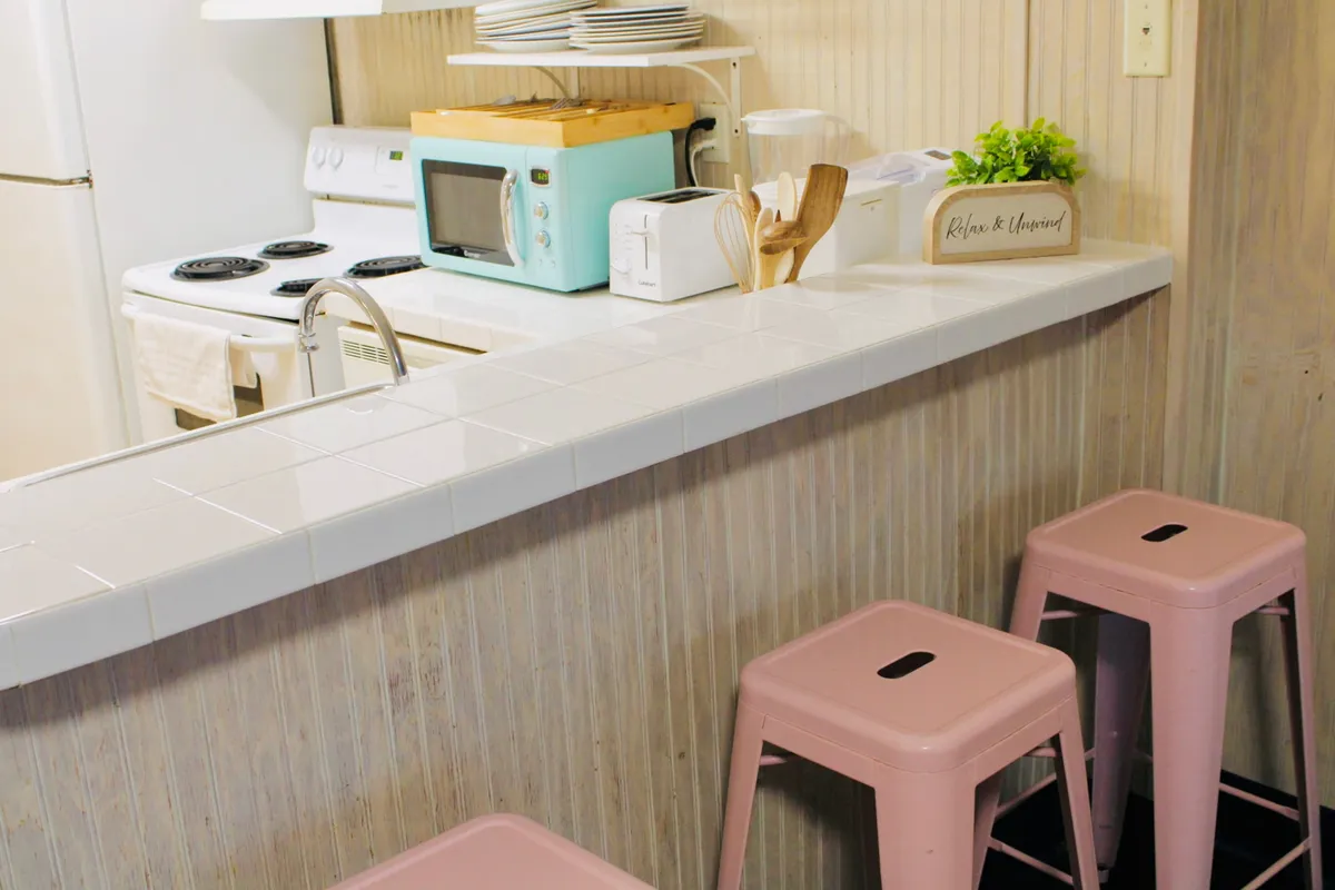 This image presents a cheerful, light-filled kitchen and dining nook with pink stools, a turquoise microwave, and white countertops. The scene invites moments of morning coffee, casual breakfasts, and socialization, reflecting a bright, coastal atmosphere.