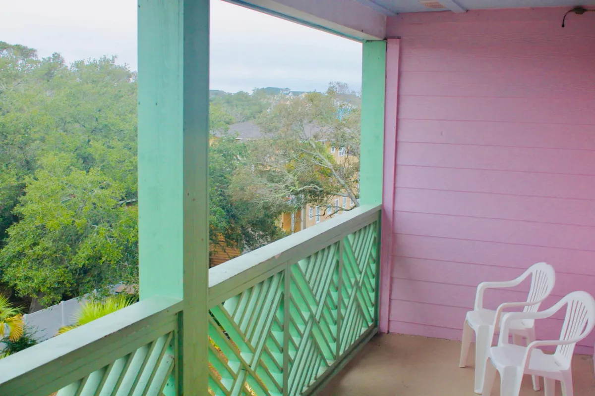 An exterior view of a pink-walled balcony framed by a green railing, with two white chairs overlooking lush greenery. The space feels inviting for morning coffee, quiet moments, or simply enjoying the outdoors with a pet.