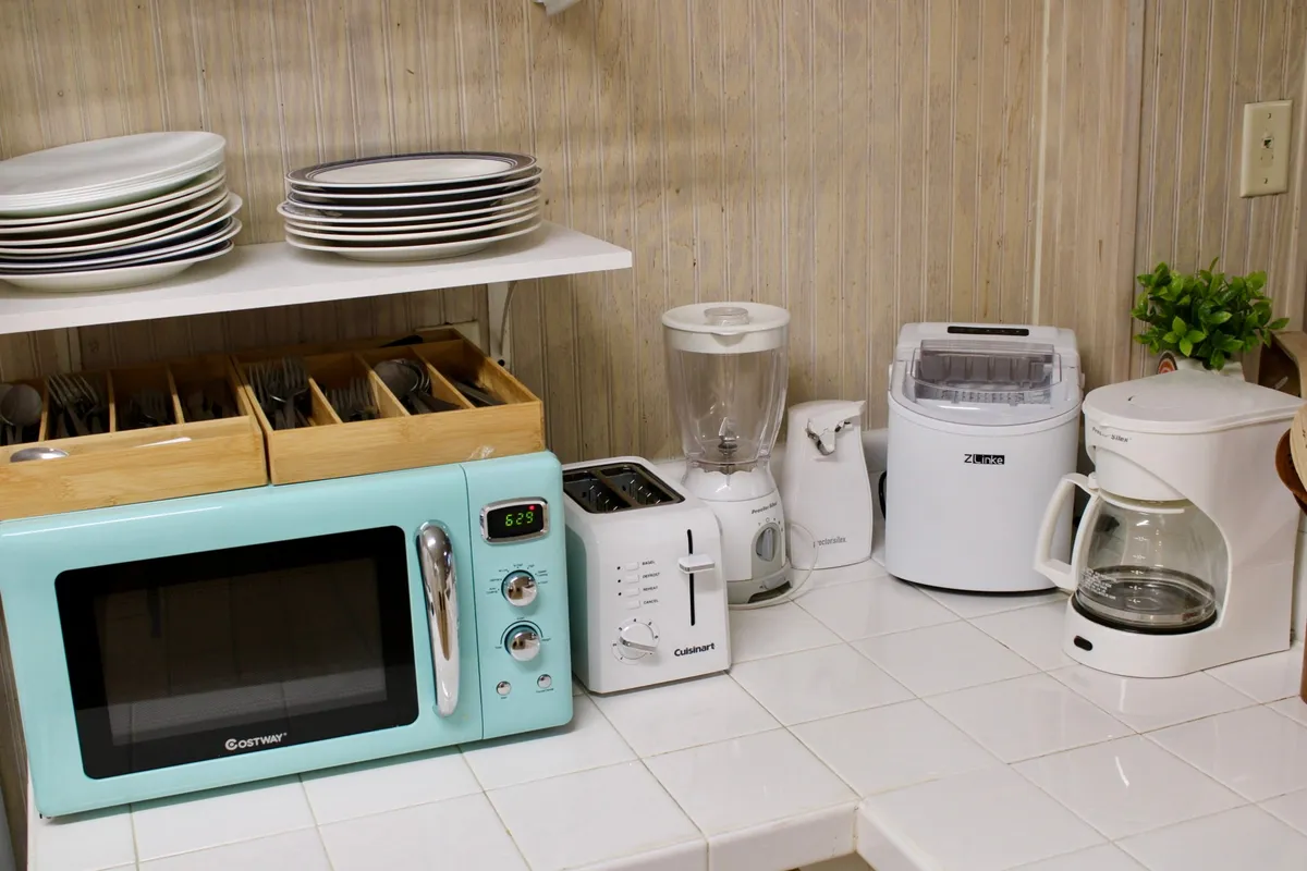 A compact kitchenette featuring a mint green microwave, a sleek white coffeemaker, a toaster, a blender, and a set of neatly arranged dishware. The setup exudes a cozy, efficient vibe ideal for preparing light meals and morning beverages.